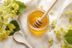 Pot de miel de tilleul doré avec fleurs de tilleul et cuillère en bois, illustrant les bienfaits du miel de tilleul